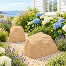 Two tan artificial landscape rocks in a coastal cottage garden, surrounded by light decorative gravel, blue and white hydrangeas, lavender, and ornamental grasses beside a white house overlooking the ocean.