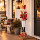 Red and gold Christmas nutcracker king holding a gold scepter, displayed on a covered front porch beside a black door, holiday wreath, lantern, and festive greenery.
