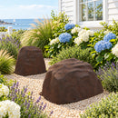 Two brown artificial landscape rocks in a coastal cottage garden, surrounded by decorative gravel, blue and white hydrangeas, lavender, and ornamental grasses beside a white house with an ocean view.