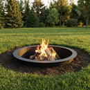Black circular metal fire pit with burning logs, set into a grassy backyard surrounded by trees.