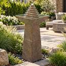Outdoor pedestal fountain with a tiered pyramid water feature and square basin, displayed in a landscaped patio garden with ornamental grasses and shrubs.