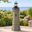 Weathered brown and green lighthouse statue displayed on a stone patio surrounded by green shrubs, flowering plants, ornamental grasses, and a coastal view in the background.