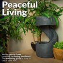Dark gray indoor tabletop fountain with a curved cascading design, shown among green plants and creating a gentle, calming water flow.