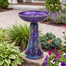 Crackled Glass Mosaic Bird Bath in iridescent purple shown outdoors in a landscaped garden, featuring a shimmering purple and multicolor crackled glass bowl and tapered pedestal surrounded by flowers and greenery.
