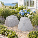 Two light gray artificial landscape rocks with realistic stone textures displayed side by side in a coastal garden with decorative pebbles, flowering hydrangeas, ornamental grasses, and a white house overlooking the water.