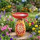 Red Mediterranean-style glass bird bath with a vibrant red, orange, green, and cream patterned pedestal, surrounded by colorful flowers, with two birds near the water.