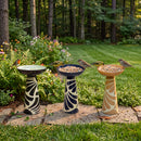 Three decorative pedestal bird baths in black, blue, and tan with cream patterns, displayed in a garden with birds perched on the rims and surrounded by flowers and greenery.