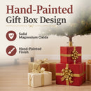Present Pile Decorative Christmas Tree Base shown beneath an artificial Christmas tree, featuring solid magnesium oxide construction and a hand-painted stacked red gift box design with gold ribbon and bow details.