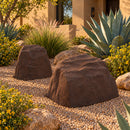 Two brown artificial landscape rocks in a Southwestern desert garden, surrounded by tan decorative gravel, agave plants, ornamental grasses, yellow flowering shrubs, and natural boulders outside a stucco home.