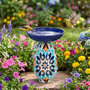 Colorful Mediterranean-style glass bird bath with an intricate blue, orange, and cream patterned pedestal, surrounded by vibrant flowers, with two birds flying and perched near the water.