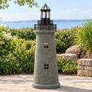 Weathered brown and green lighthouse statue displayed on a stone patio surrounded by green shrubs, flowering plants, ornamental grasses, and a coastal view in the background.