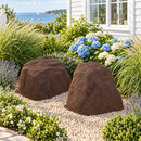 Two dark brown artificial landscape rocks with realistic stone textures displayed side by side in a coastal garden with decorative pebbles, blue hydrangeas, flowering plants, and ornamental grasses beside a white house overlooking the water.