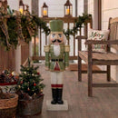 Green and red Christmas nutcracker statue displayed on a covered porch with holiday garland, potted greenery, lanterns, and a rustic wooden chair.