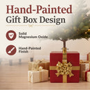 Pretty Present Decorative Christmas Tree Base shown beneath an artificial Christmas tree, featuring solid magnesium oxide construction and a hand-painted red gift box design with gold ribbon and bow details.