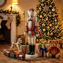 Red and green Christmas nutcracker king holding a gold scepter, displayed beside a decorated Christmas tree, wrapped gifts, a teddy bear, and toy train in a cozy holiday living room.