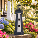 Navy and white striped lighthouse statue displayed on a brick garden wall surrounded by blue hydrangeas, pink flowers, and greenery in a landscaped front yard.