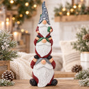 Stacked Christmas gnome statue displayed on a table in a cozy holiday setting with warm lights, winter greenery, pinecones, and a fireplace in the background.