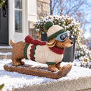 Winter dachshund statue wearing a knit hat, goggles, sweater, and scarf displayed outdoors on a snow-covered porch.