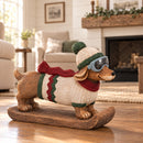 Winter dachshund statue wearing a knit hat, goggles, sweater, and scarf displayed indoors beside a fireplace.