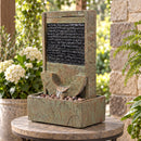 Outdoor wall fountain with a textured rectangular water feature, curved water spillway, decorative rocks, and lower reservoir, displayed on a patio table surrounded by plants.