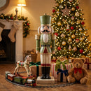 Green and red Christmas nutcracker statue displayed beside a decorated Christmas tree, wrapped gifts, a teddy bear, and toy train in a cozy holiday living room.