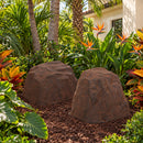 Two dark brown artificial landscape rocks with realistic stone textures displayed side by side in a tropical garden, surrounded by palm trees, lush greenery, colorful foliage, and bird-of-paradise flowers beside a stucco home.