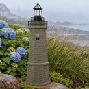 Weathered brown and green lighthouse statue displayed on a rocky coastal landscape with blue hydrangeas, ornamental grasses, and an ocean view in the background.
