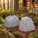 Two light gray artificial landscape rocks with realistic stone textures displayed side by side in a rustic woodland garden beside a log cabin, surrounded by ferns, moss-covered stones, and mulch in warm afternoon sunlight.