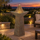 Outdoor pedestal fountain with a tiered pyramid water feature and illuminated water, displayed on a landscaped patio at sunset.