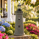 Weathered brown and green lighthouse statue displayed on a brick garden wall surrounded by blue hydrangeas, pink flowers, and greenery in a landscaped front yard.