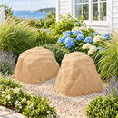 Two tan artificial landscape rocks with realistic sandstone textures displayed side by side in a coastal garden with decorative pebbles, blue hydrangeas, flowering plants, and ornamental grasses beside a white house overlooking the water.