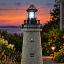 Weathered brown and green lighthouse statue with an illuminated lantern displayed at dusk overlooking a coastal landscape.